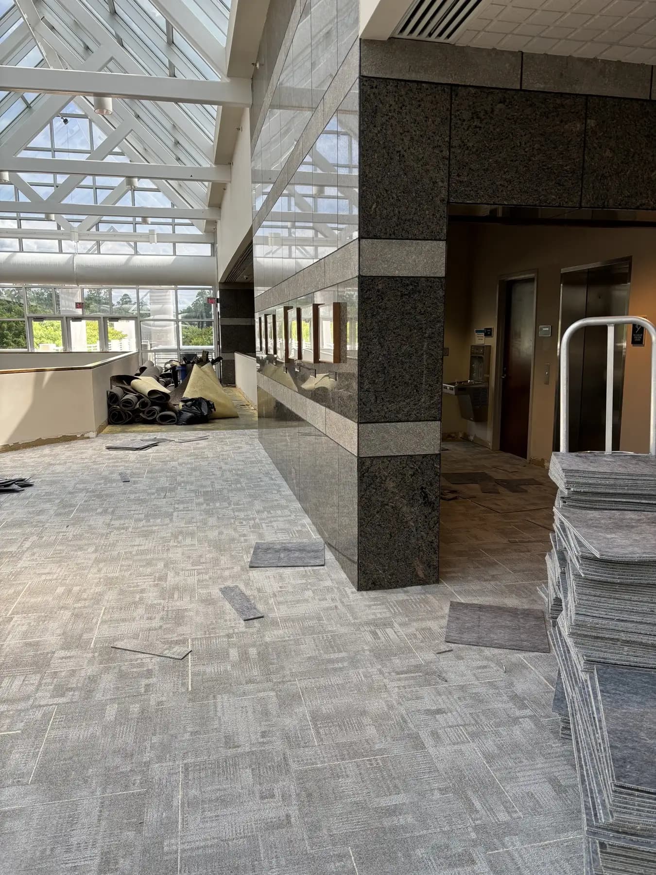 Gray commercial carpet tile laid across the atrium beside a granite elevator core under a skylight
